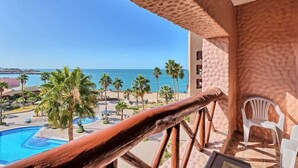 A balcony with a view of a pool and palm trees, a wooden railing, and a white plastic chair. - Charming Condo in Great Location Pinacate 412 (Puerto Peñasco)