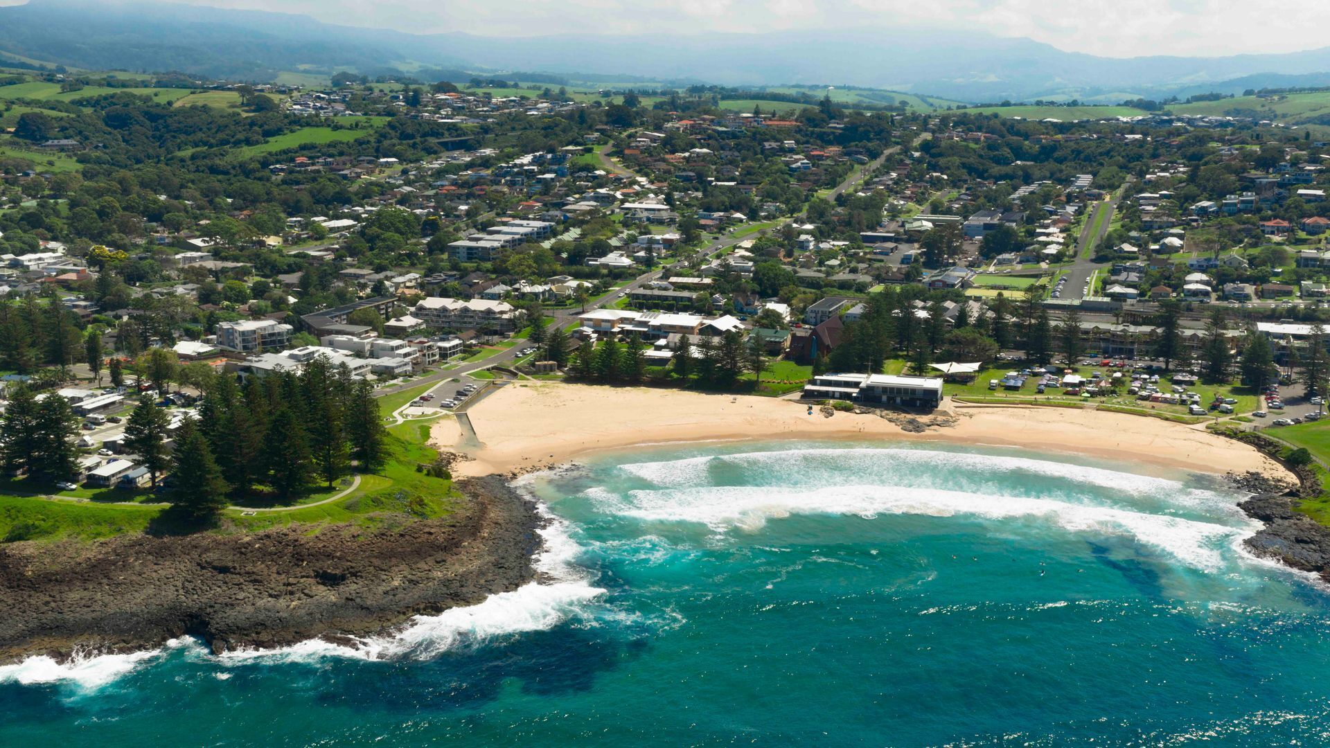 An aerial view of a coastal town with a sandy beach, a rocky shoreline, and a mix of residential and commercial buildings.