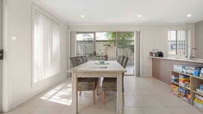 A modern kitchen with a dining area, featuring a large table, wicker chairs, and a variety of board games on shelves.