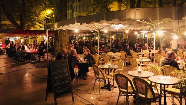 Een terras buiten met tafels en stoelen onder een overkapping, omgeven door bomen en gebouwen.