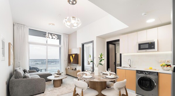 Ein modernes Wohnzimmer mit einer Couch, einem Esstisch und Stühlen, eine Küche mit Mikrowelle, Spüle und Waschmaschine, und ein großes Fenster mit Blick.