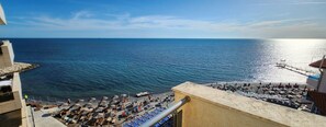 Ein Strand mit Sonnenschirmen und Liegestühlen, einer Mole und freier Sicht auf das Meer.