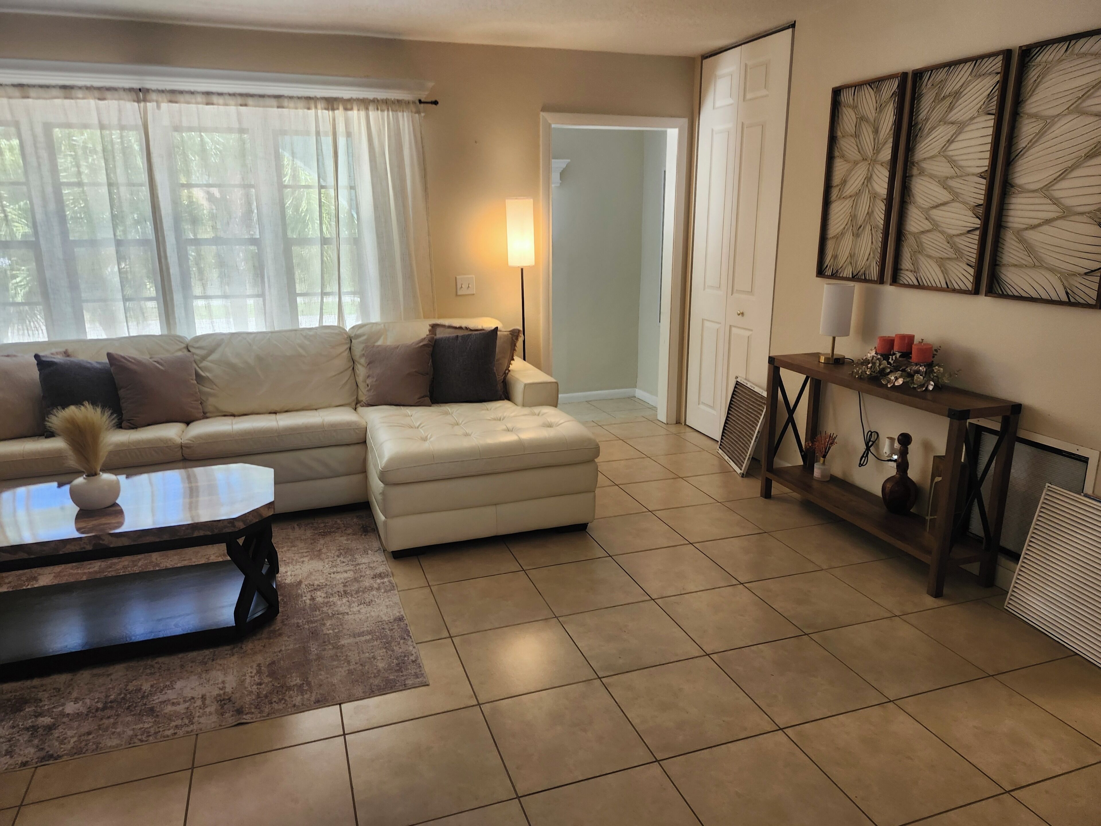 A living room with a sofa, ottoman, coffee table, and a side table with decorative items. There is a door leading to another room and a wall with framed artwork.