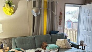 A cozy living room with a sectional sofa, a small table with a lamp, and a view of the outside through a door. - Nature At the Cozy House (Slidell)
