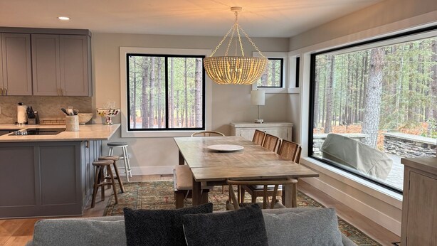 Une cuisine moderne avec des meubles gris, un coin repas avec une table et des chaises en bois, et une grande fenêtre donnant sur la forêt.