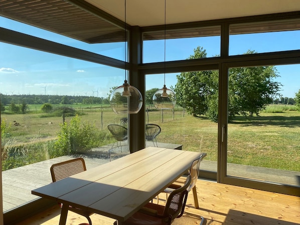A modern dining area with a large table and chairs, large windows offering a view of a grassy field and wind turbines.
