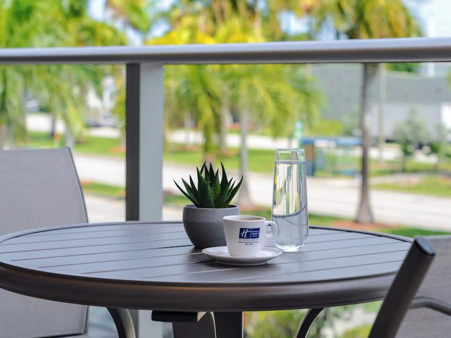 Ein runder Tisch mit einer weißen Tasse, einem Glas Wasser und einem Blumentopf auf einem Balkon mit Blick auf eine Straße mit Palmen.