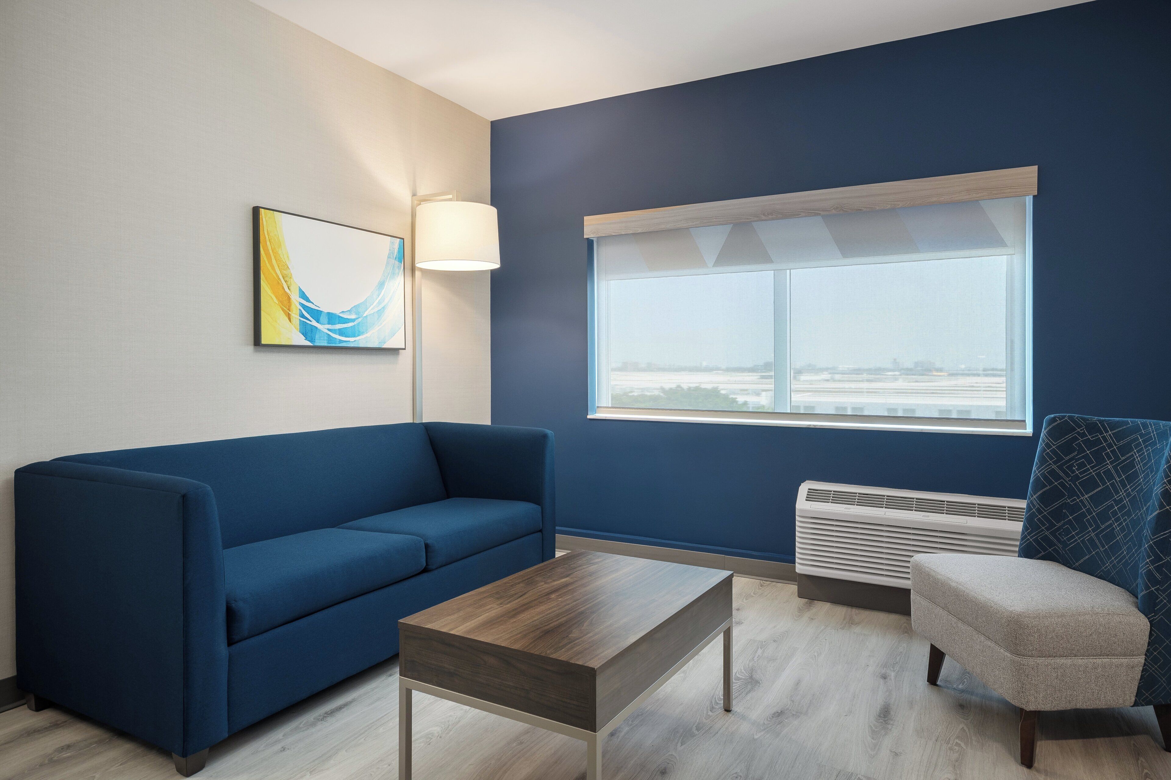 A room with a blue sofa, a wooden coffee table, a chair with a geometric pattern, a floor lamp, and a window with a view.