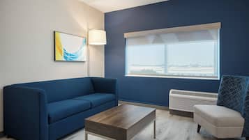 A room with a blue sofa, a wooden coffee table, a chair with a geometric pattern, a floor lamp, and a window with a view.