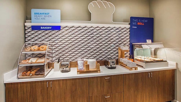 A bakery counter with a sign indicating breakfast is ready, a display case with pastries, a sign about how they roll, and a wall with a geometric pattern. - Holiday Inn Express & Suites Miami Intl Airport - 36th St (Miami)