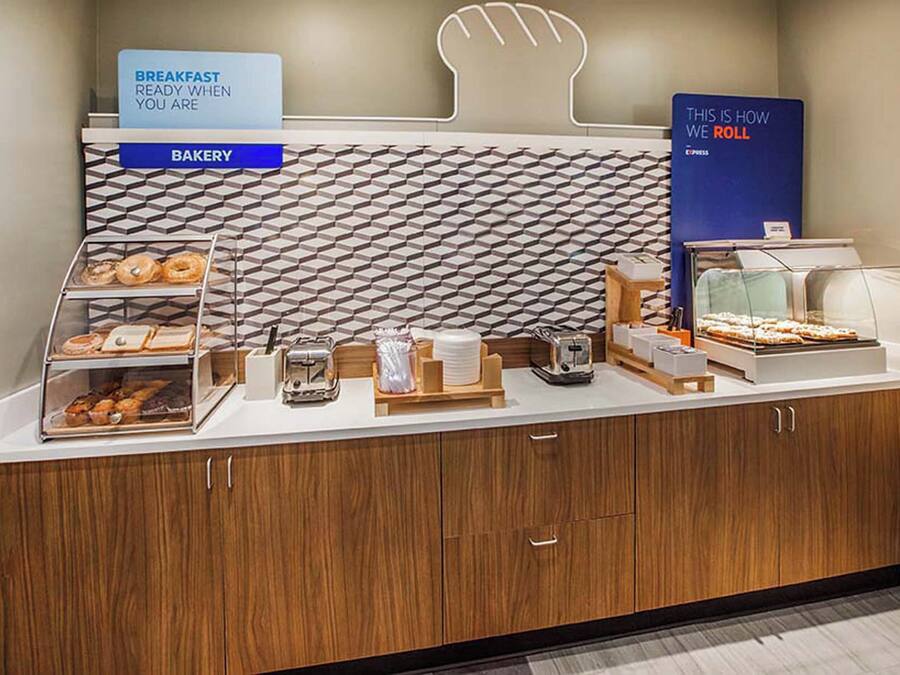 A bakery counter with a sign indicating breakfast is ready, a display case with pastries, a sign about how they roll, and a wall with a geometric pattern.