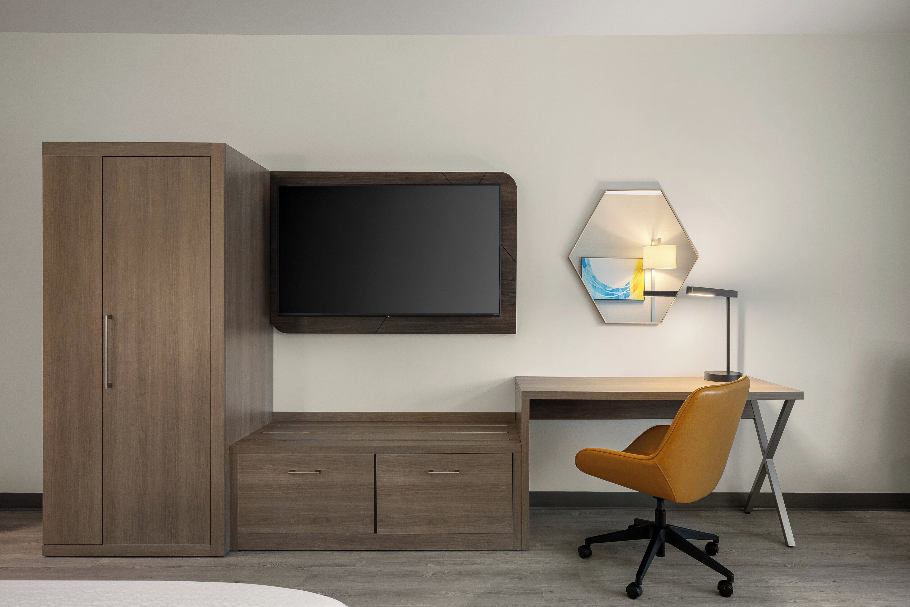 A modern room with a wooden wardrobe, a desk with a chair, a flat-screen TV, and a wall-mounted lamp.