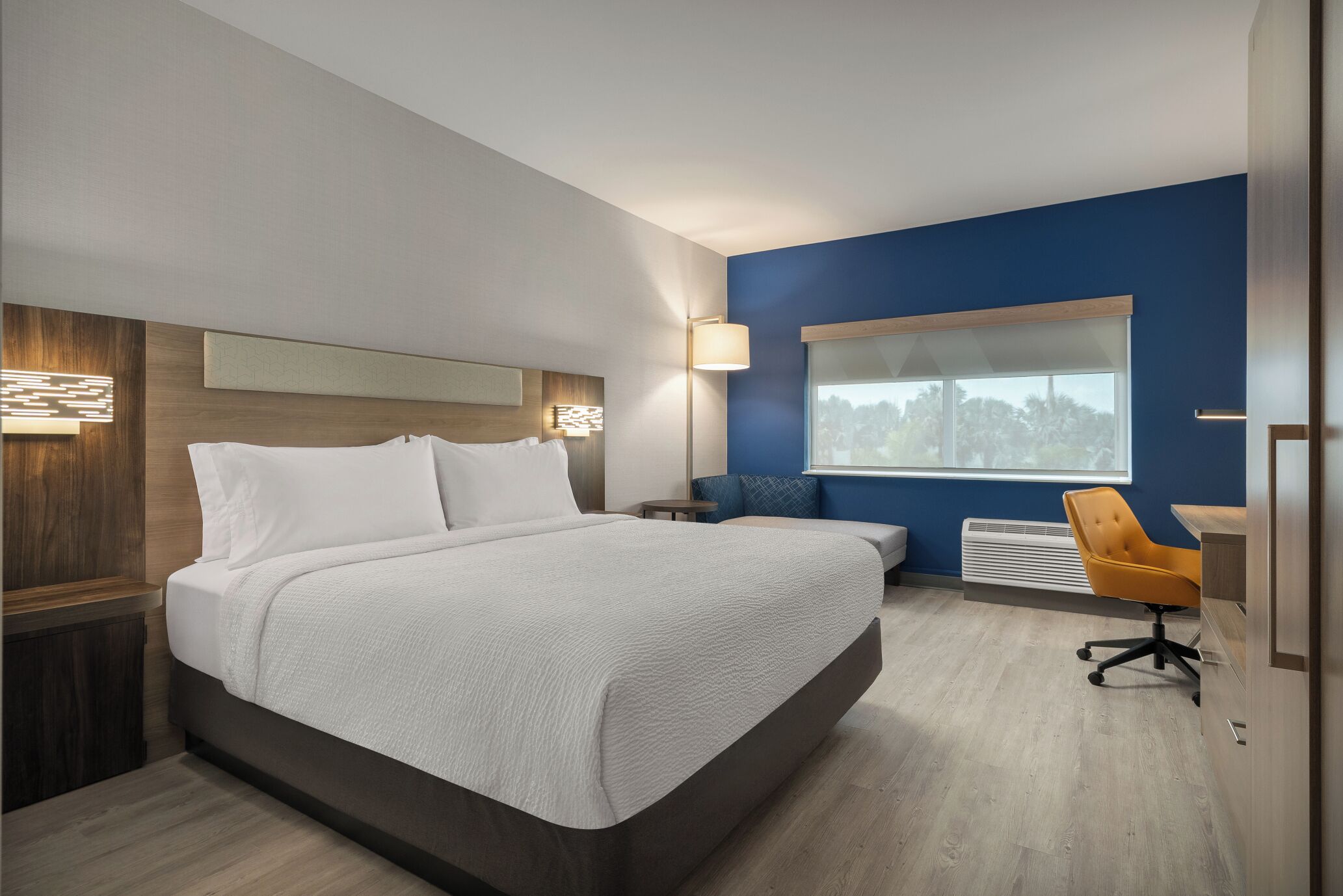 A modern hotel room with a large bed, a desk, a chair, and a window with a view.