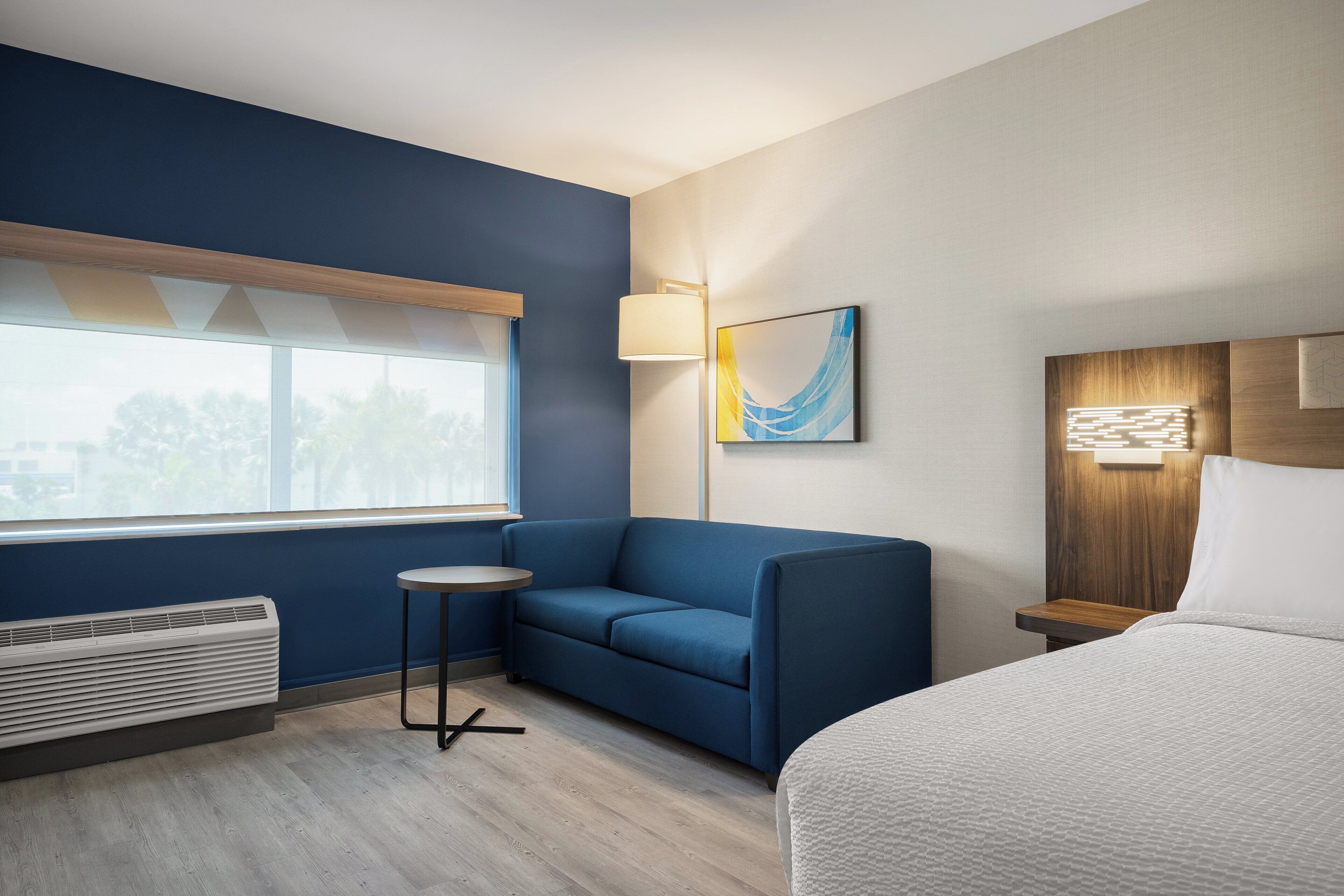 Une chambre d’hôtel avec un canapé bleu, une table ronde, un lit, une fenêtre avec des stores, une lampe fixée au mur et un tableau.