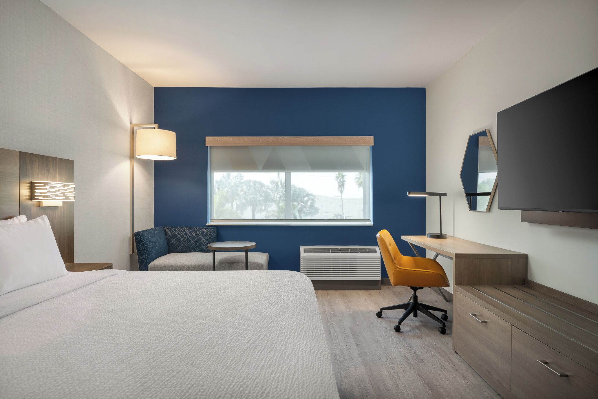 A hotel room with a bed, a desk, a chair, a window, a heater, and a flat-screen TV.