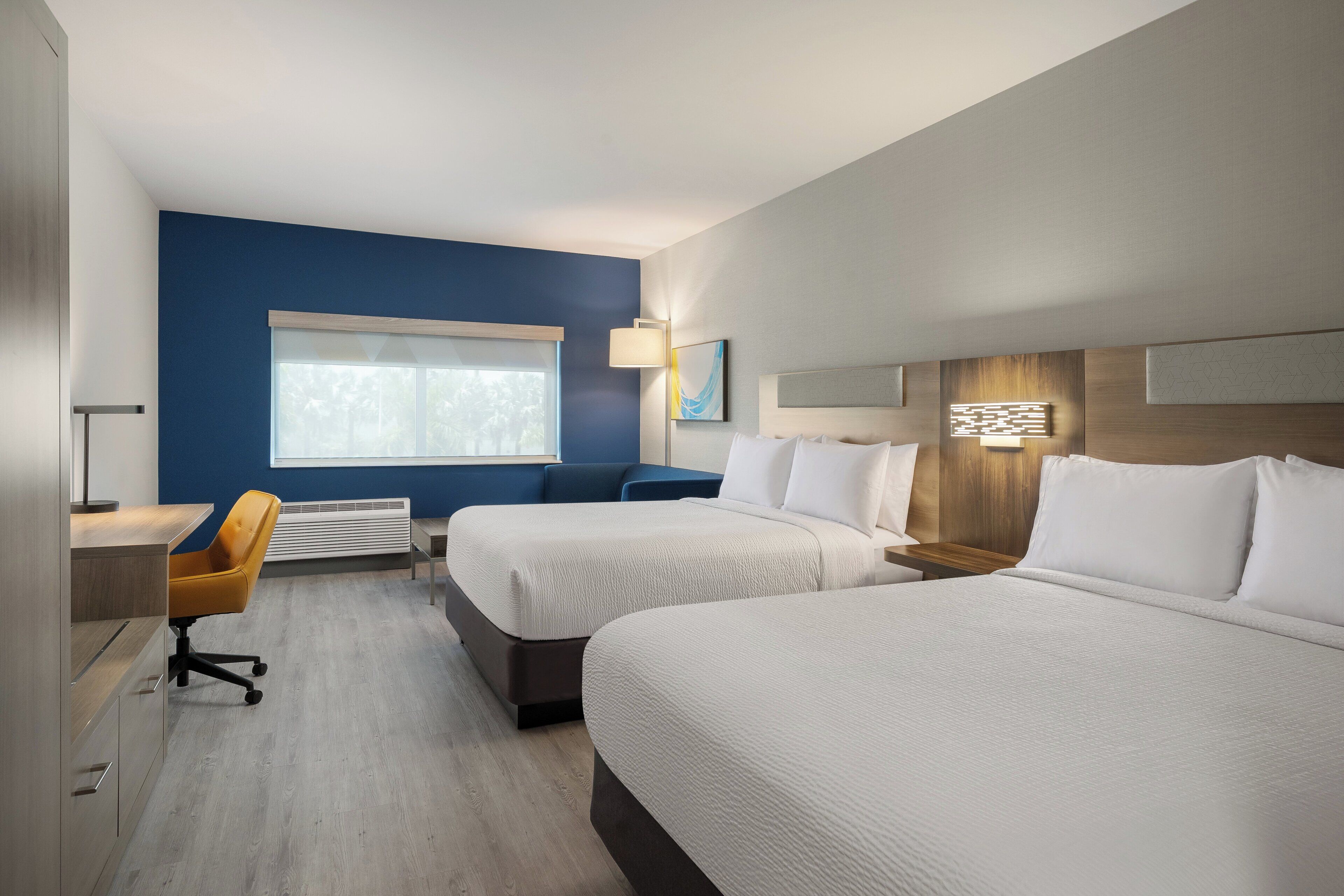 Une chambre d’hôtel avec deux lits, un bureau, une chaise, une fenêtre et une lampe fixée au mur.