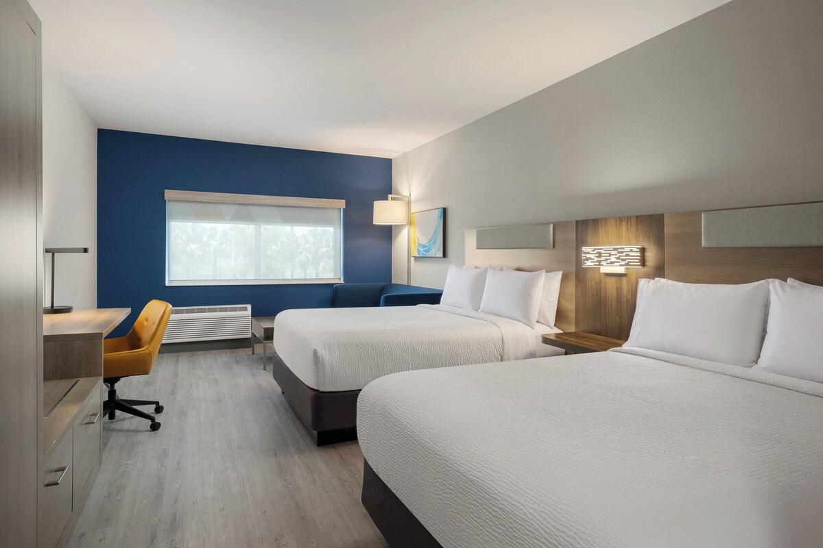 Ein Hotelzimmer mit zwei Betten, einem Schreibtisch, einem Stuhl, einem Fenster und einer Wandlampe.