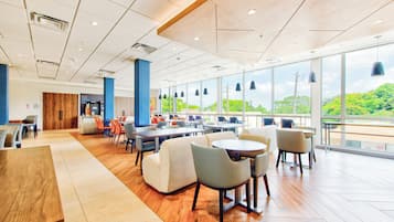 A spacious indoor area with multiple seating arrangements, large windows offering a view of greenery, and a mix of wooden and carpeted flooring.