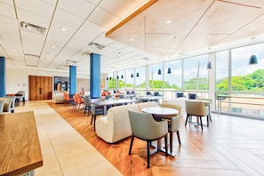 A spacious indoor area with multiple seating arrangements, large windows offering a view of greenery, and a mix of wooden and carpeted flooring. - Holiday Inn Express & Suites Miami Intl Airport - 36th St (Miami)