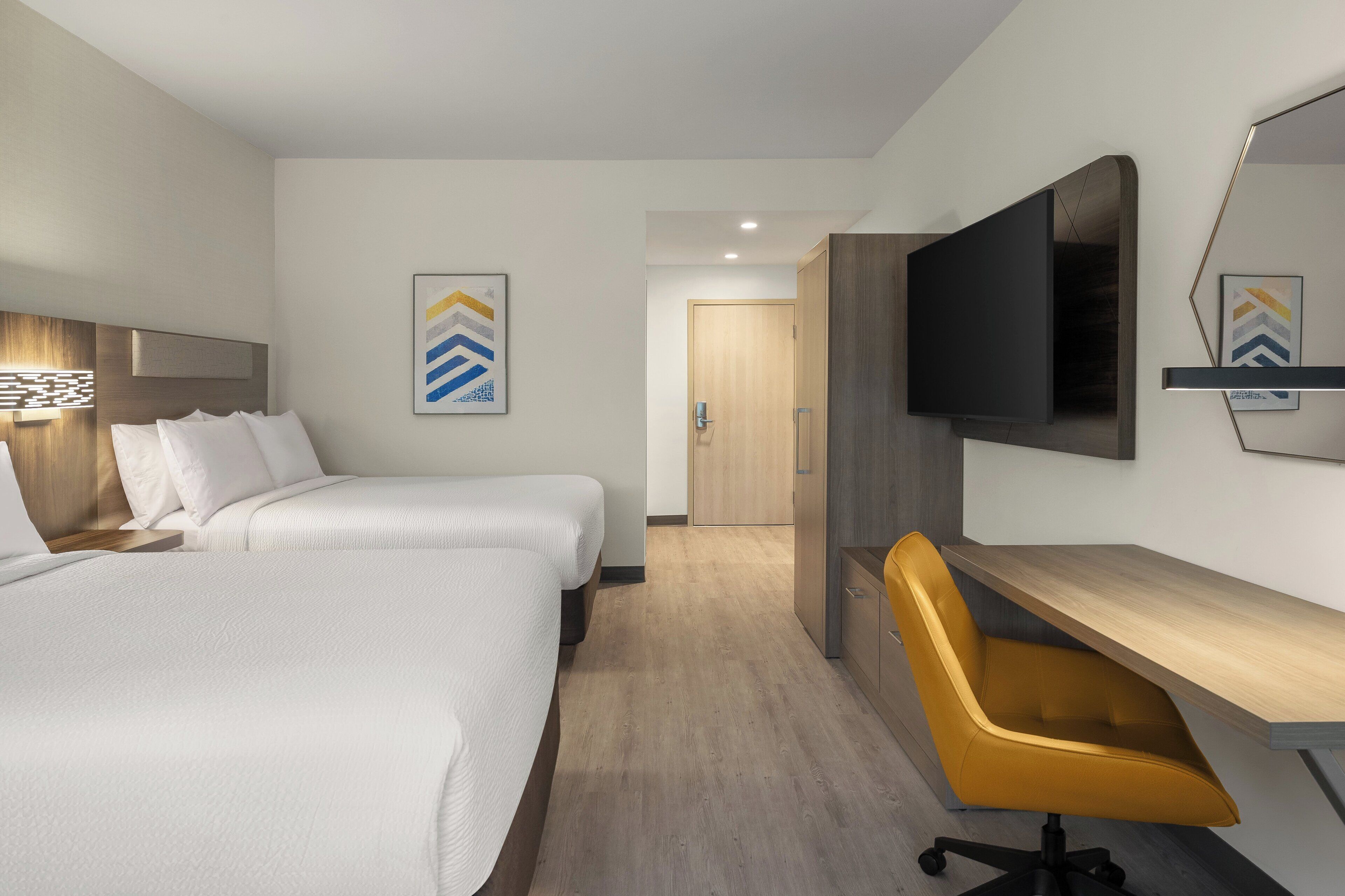 A hotel room with two beds, a desk, a chair, a flat-screen TV, and artwork on the walls.