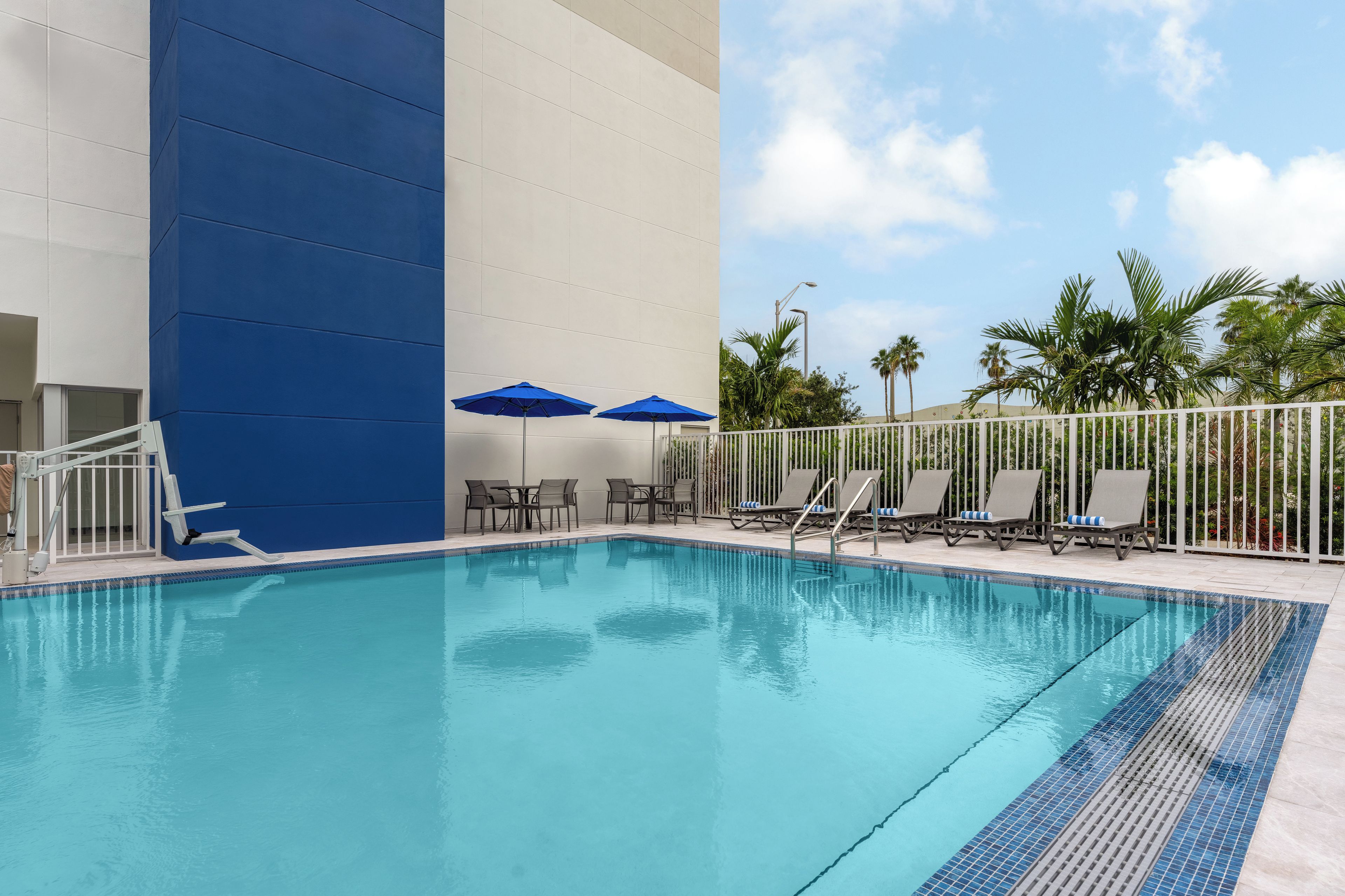 A swimming pool with lounge chairs and umbrellas, surrounded by a white fence and palm trees.