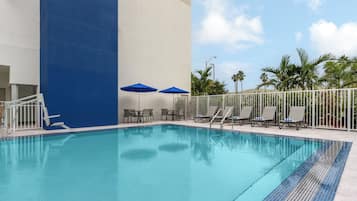 A swimming pool with lounge chairs and umbrellas, surrounded by a white fence and palm trees.