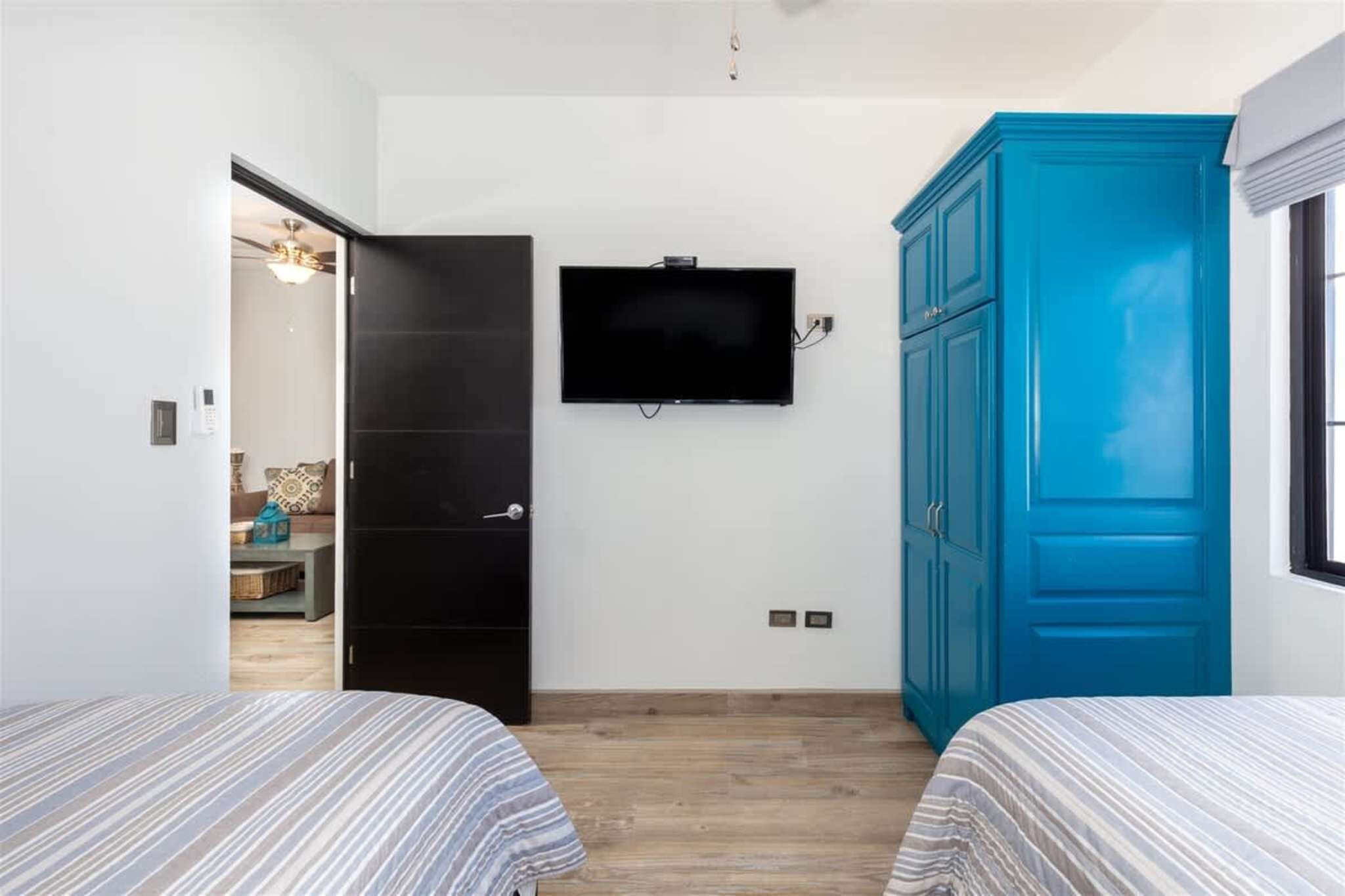 Ein Schlafzimmer mit zwei Betten, einem an der Wand montierten Fernseher, einem blauen Kleiderschrank und einer Tür zu einem anderen Raum.