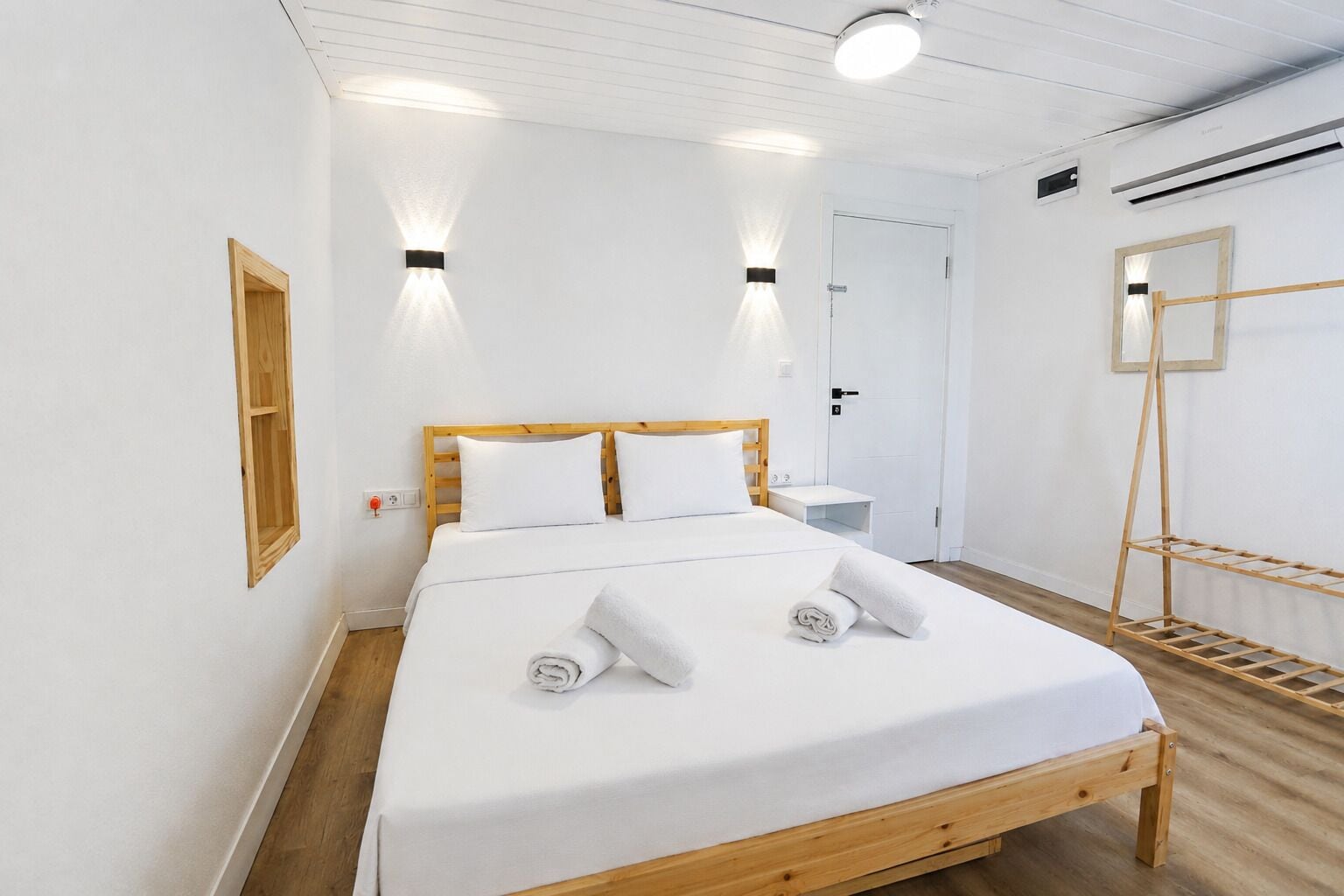 Una cama bien hecha con sábanas blancas y dos toallas enrolladas, un cabecero de madera, una mesita de noche, una lámpara fijada a la pared y un espejo.