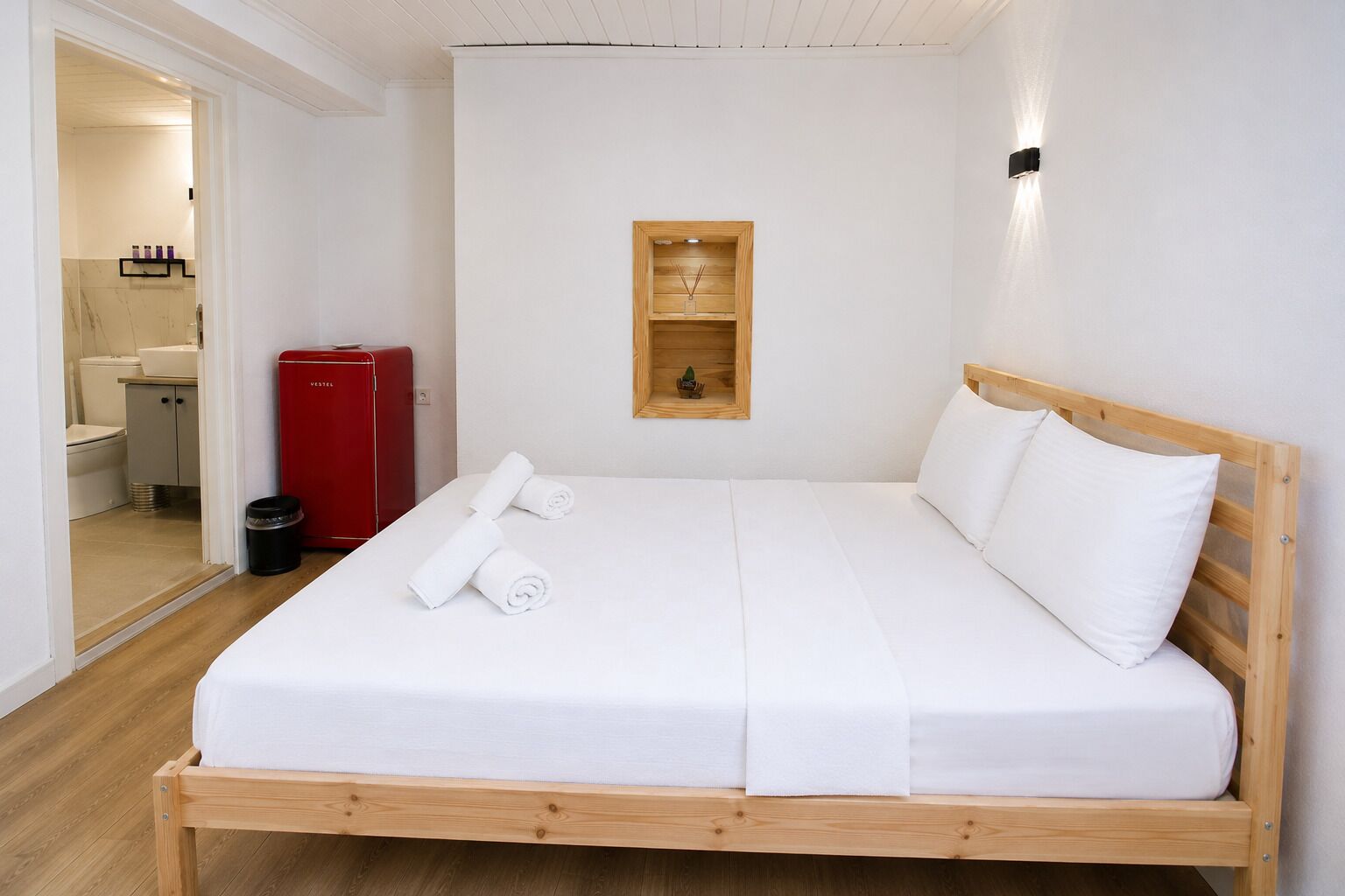 Habitación con cama individual, estructura de madera, ropa de cama blanca y una pequeña estantería de madera con un objeto decorativo.