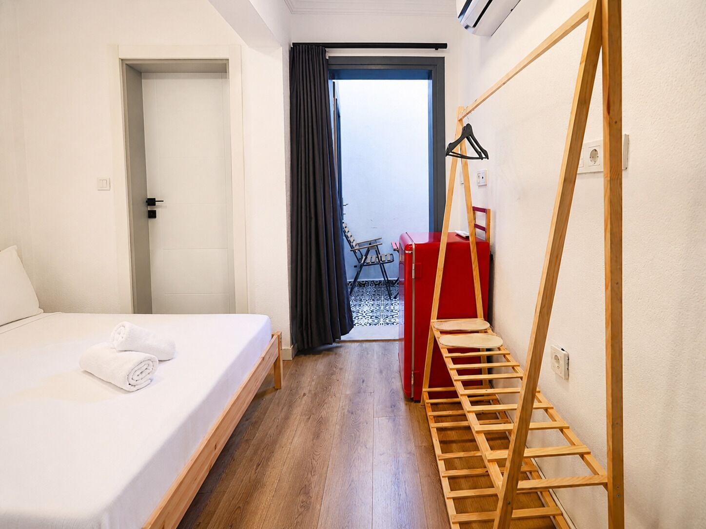 A hotel room with a bed, a wooden ladder, a red suitcase, and a view of the outside.