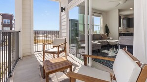 En balkon med træmøbler, en glasdør der fører til en opholdsstue, og udsigt til stranden.