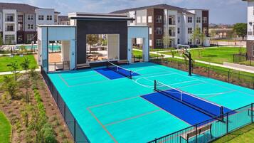Uma quadra de basquete com rede, cercada por uma grade, e próxima a edifícios residenciais.