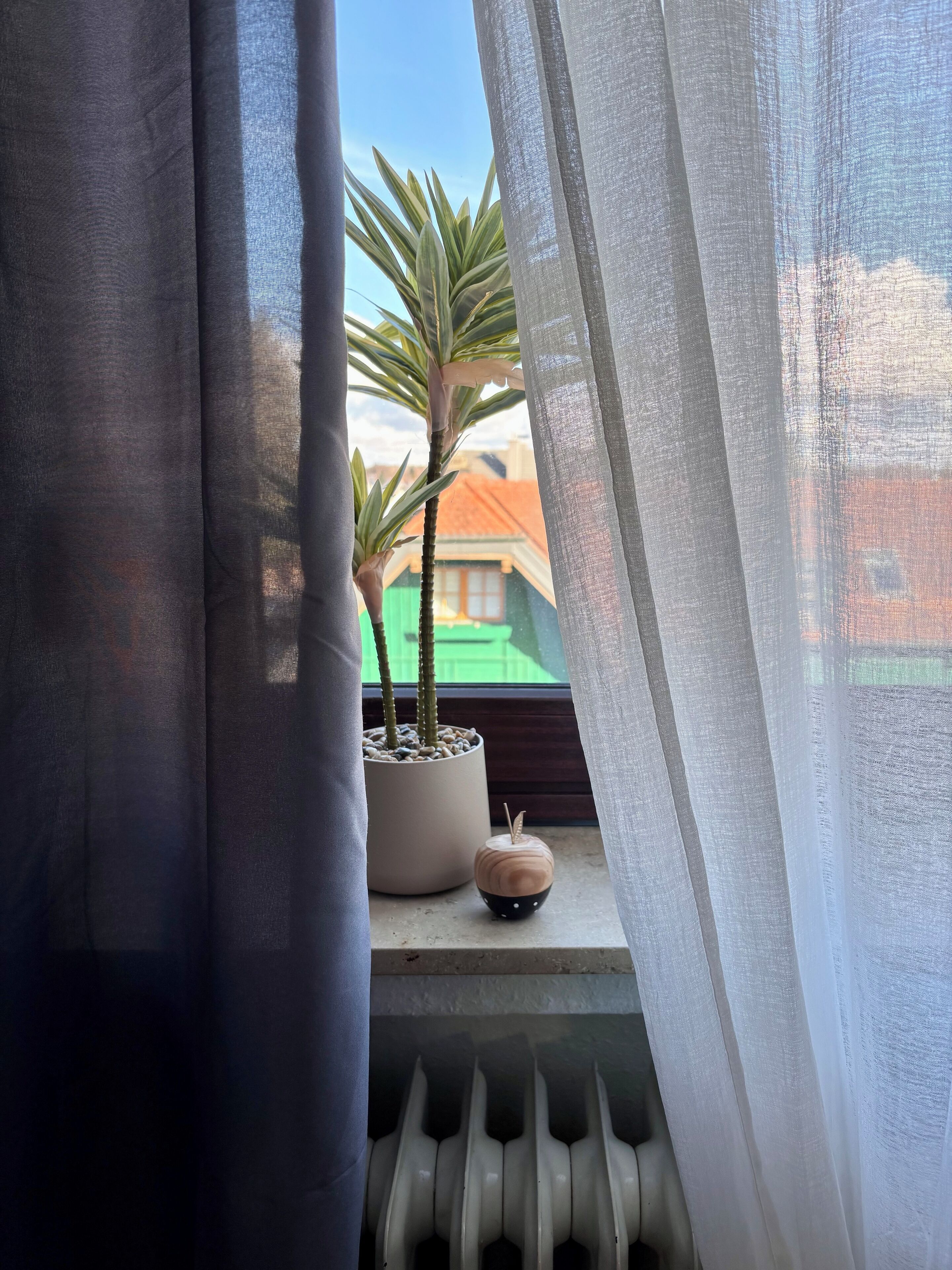 Ein Fenster mit durchsichtigen Vorhängen, einem Blumentopf und einem kleinen Holzgegenstand auf dem Fensterbrett.