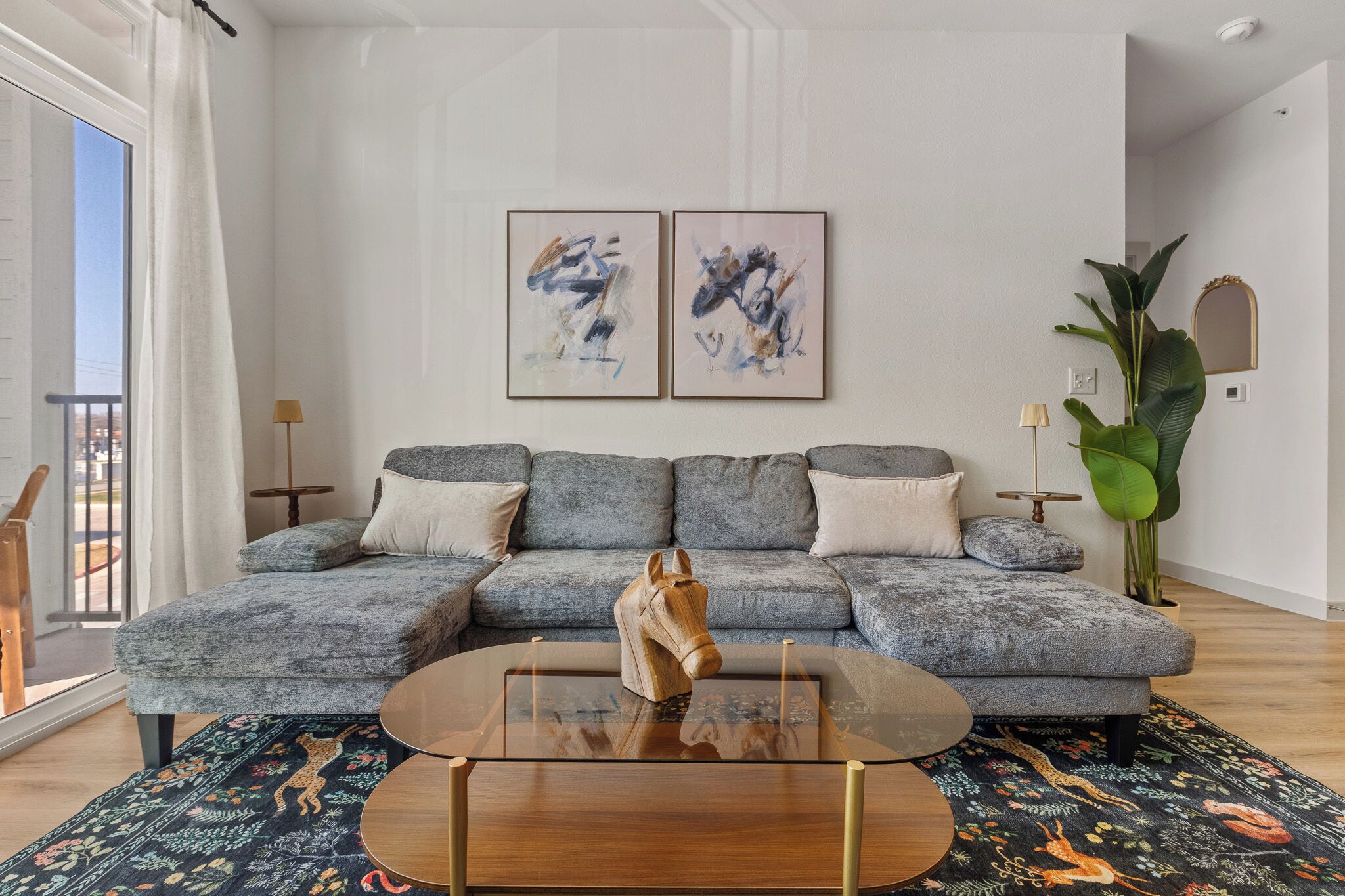 A modern living room with a grey sectional sofa, a glass coffee table with a wooden horse sculpture, abstract wall art, and a large green plant.