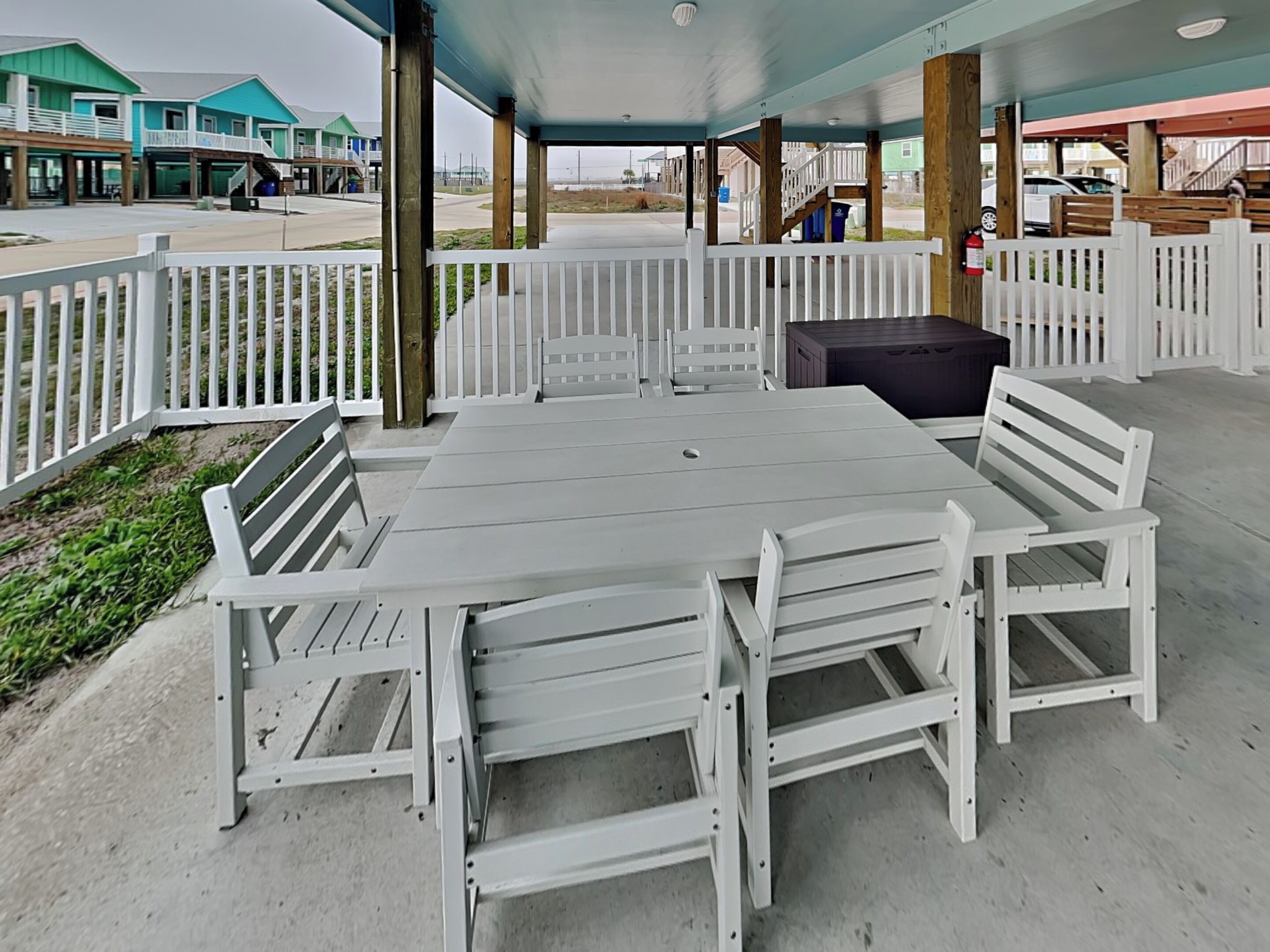 Une terrasse extérieure avec une table et des bancs, entourée d’une clôture blanche, avec des bâtiments en arrière-plan.