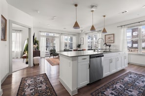 A modern kitchen with a central island, white cabinetry, stainless steel appliances, pendant lighting, and large windows offering a view of the outside. - Refined Ground Level 2BR 2BA with Patio Access (San Marcos)