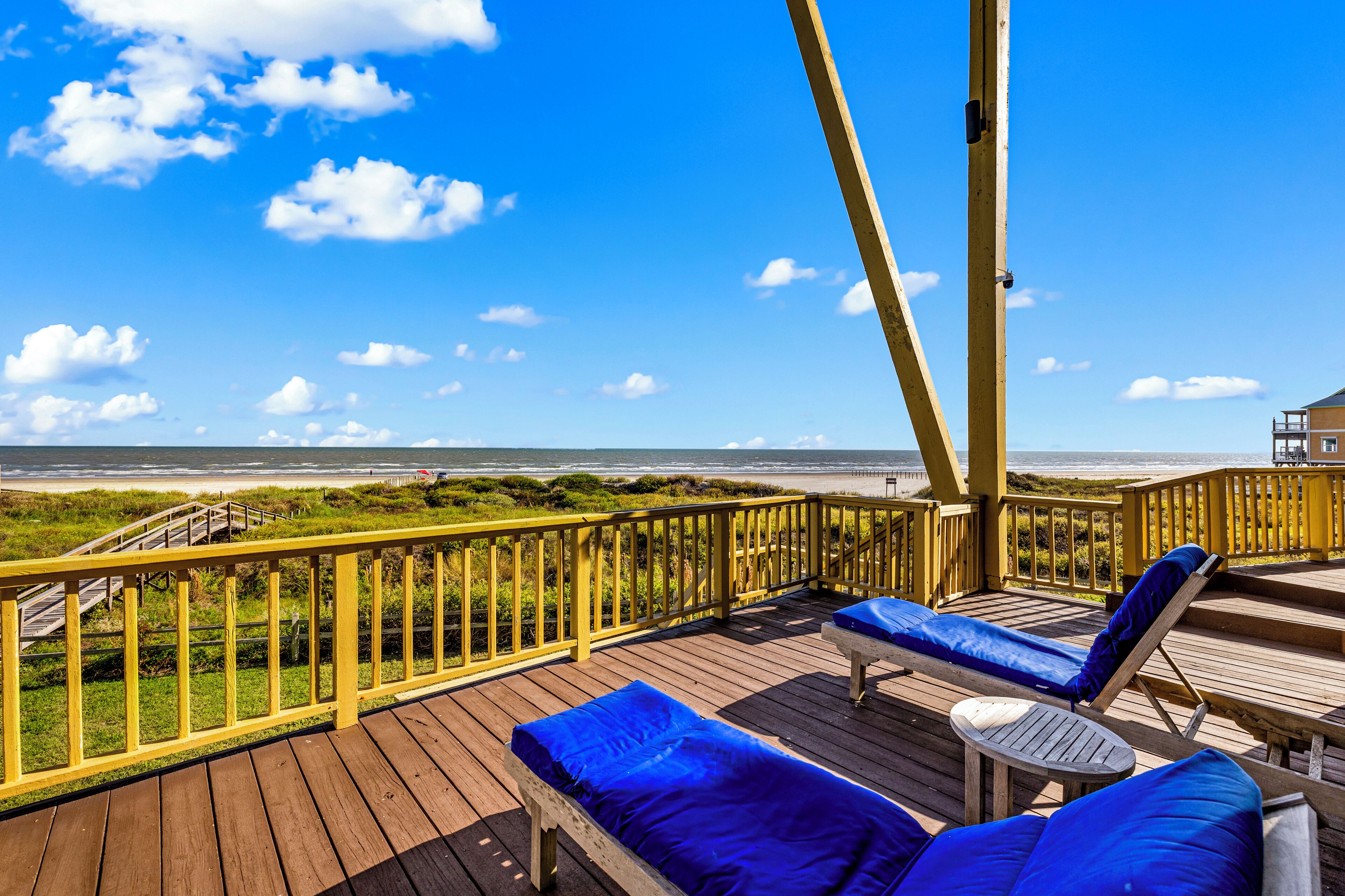A wooden deck with two lounge chairs and a small table, overlooking a beach with a wooden walkway.