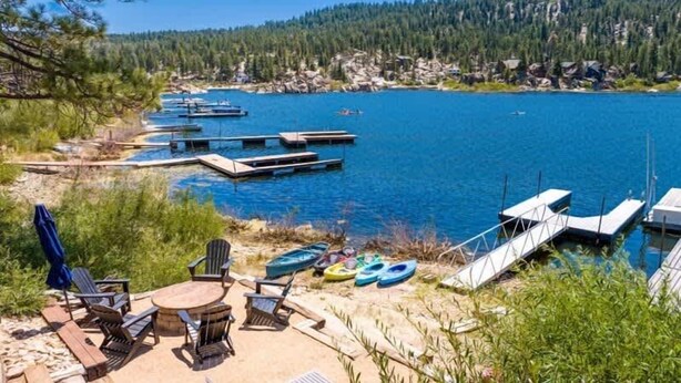A lakeside view with docked boats, a sandy area with chairs and a table, and a forested background.