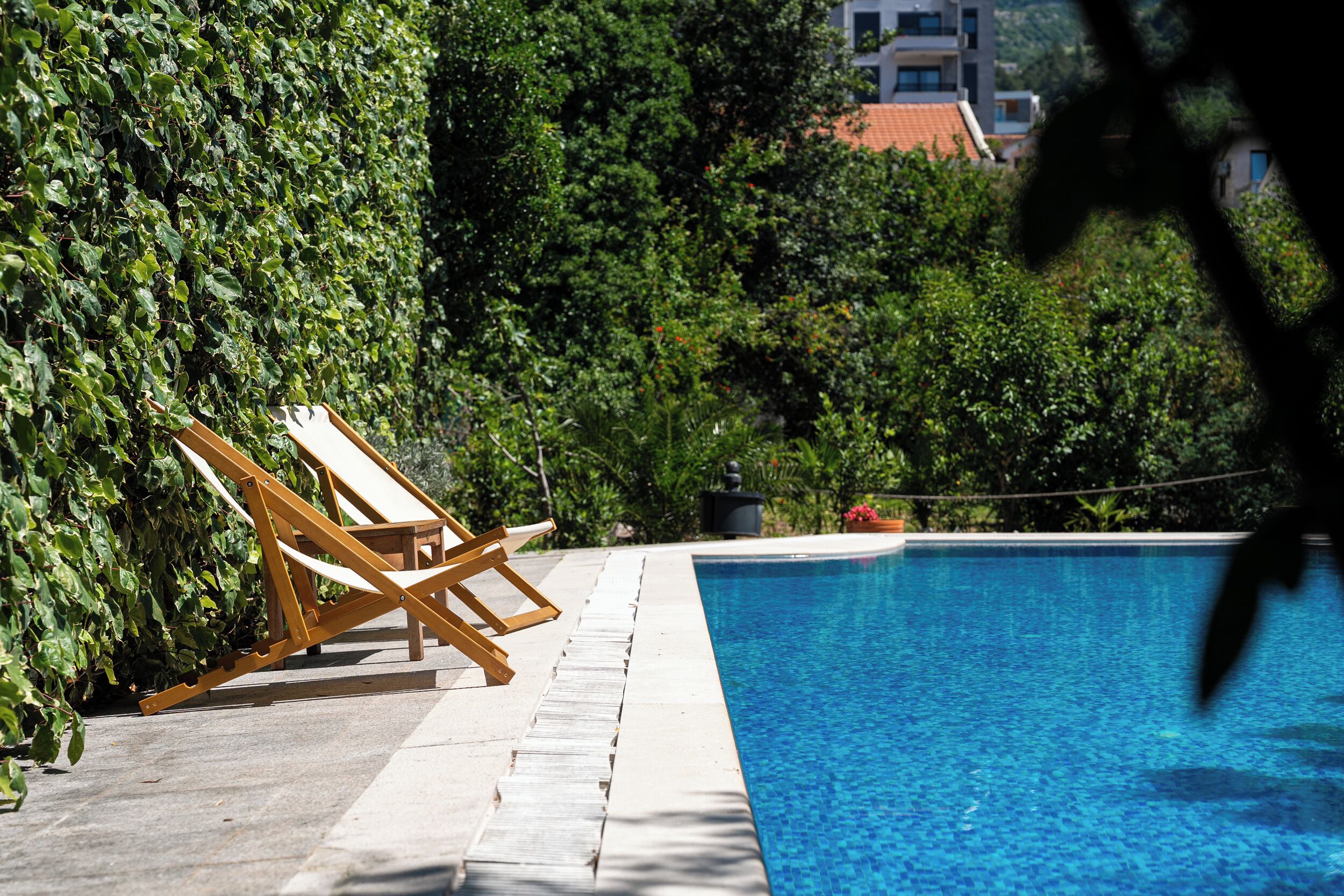 Una piscina con una tumbona de madera y un seto.