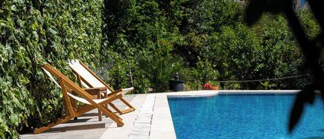 Una piscina con una tumbona de madera y un seto.