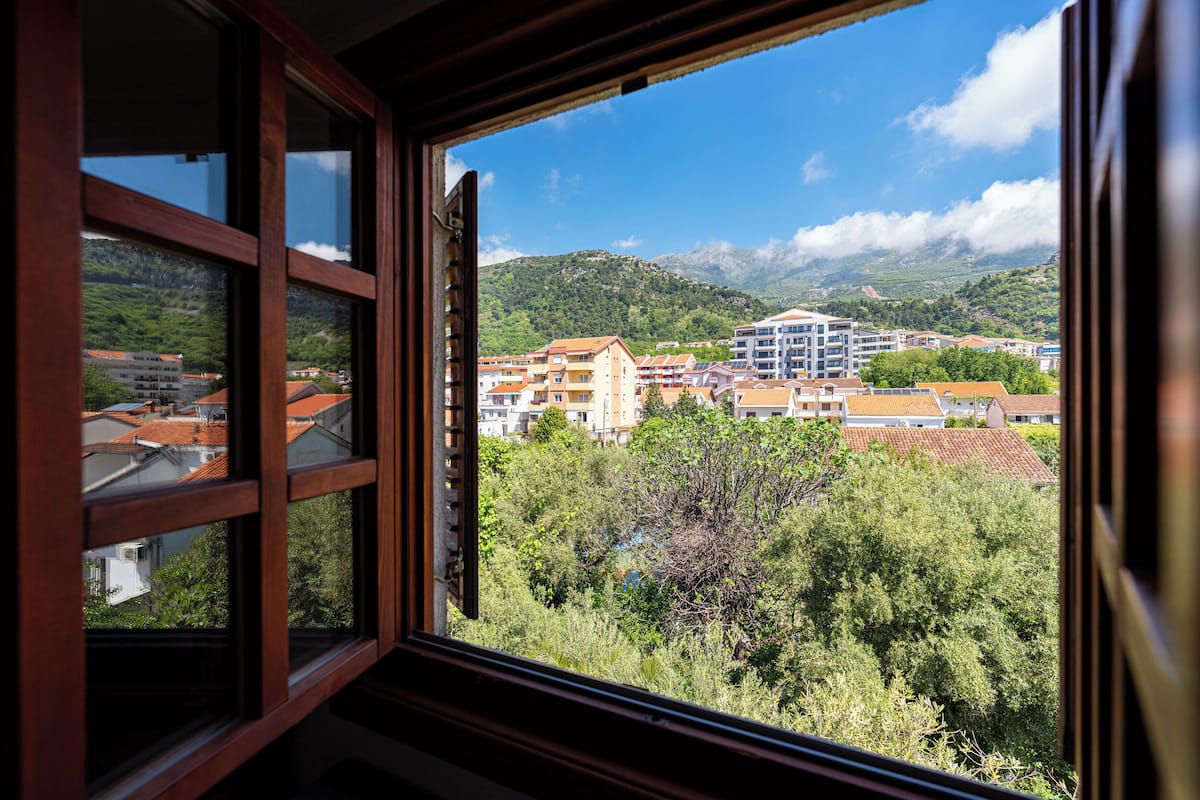 A view through a window with a wooden frame, revealing a town with buildings and greenery.