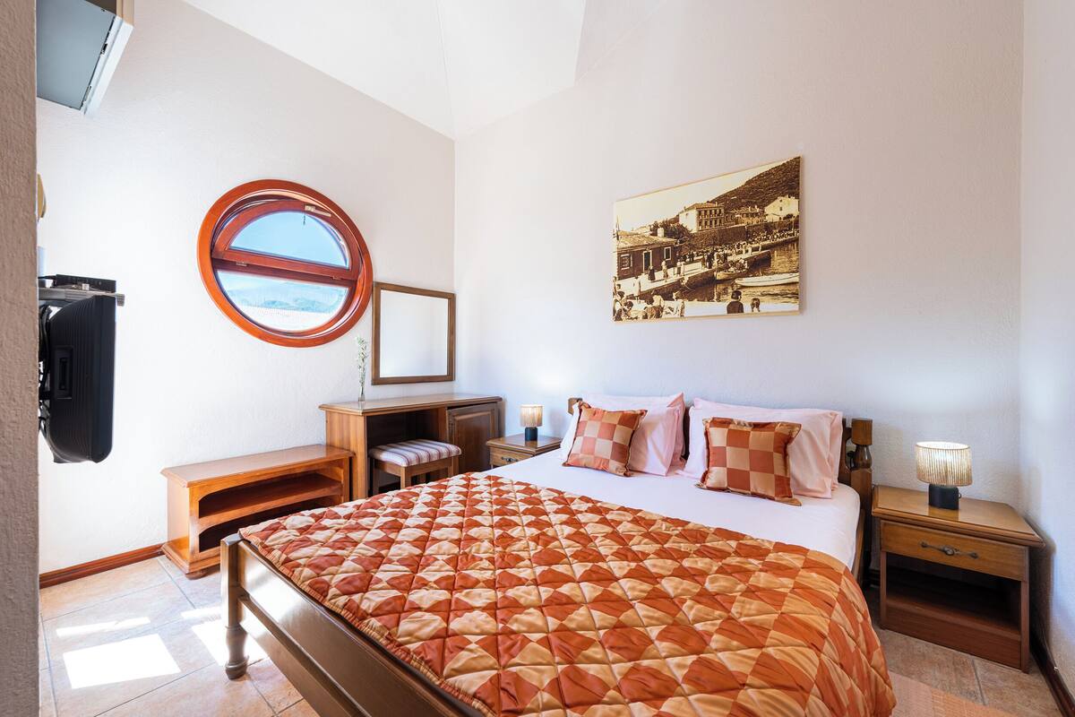 A bedroom with a bed, a round window, a bedside table, a nightstand, a lamp, a mirror, and a wall-mounted TV.