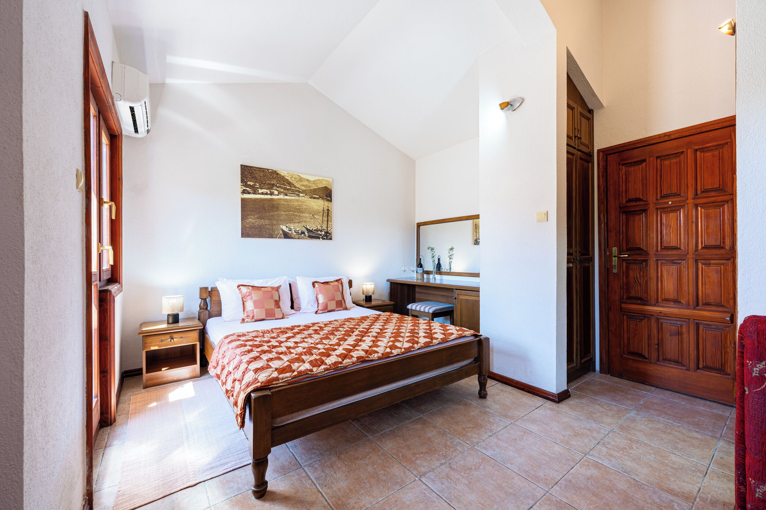 A bedroom with a bed, bedside tables, a nightstand, a wooden wardrobe, a mirror, and a painting on the wall.