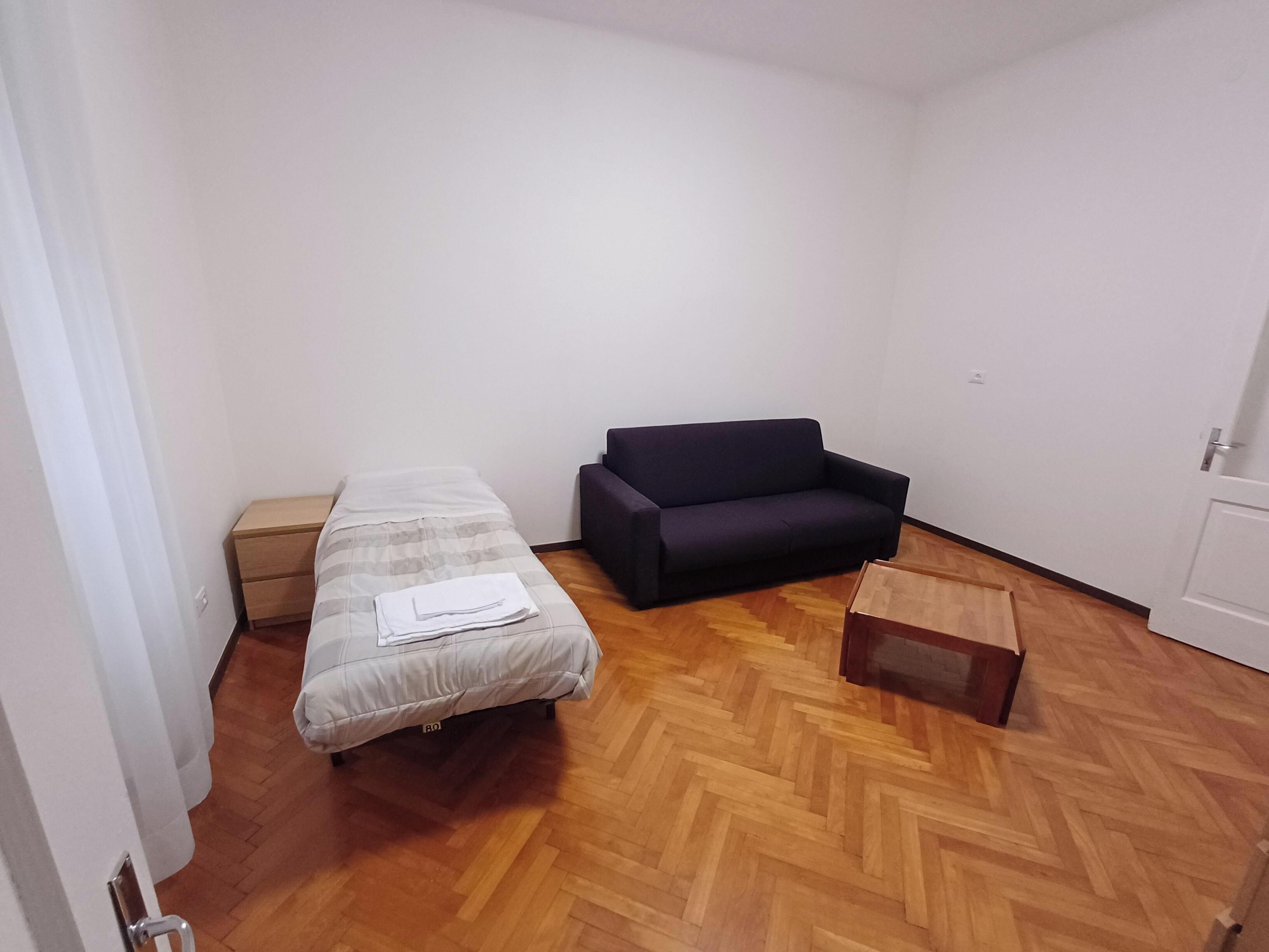 Une petite pièce, peu meublée, avec un lit, un canapé sombre, une commode en bois et une petite table en bois.