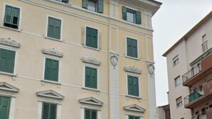 Un edifici de diverses plantes amb un estil arquitectònic clàssic, que destaca per finestres simètriques amb persianes verdes, portals d'arc i un rètol que indica un negoci a la planta baixa.