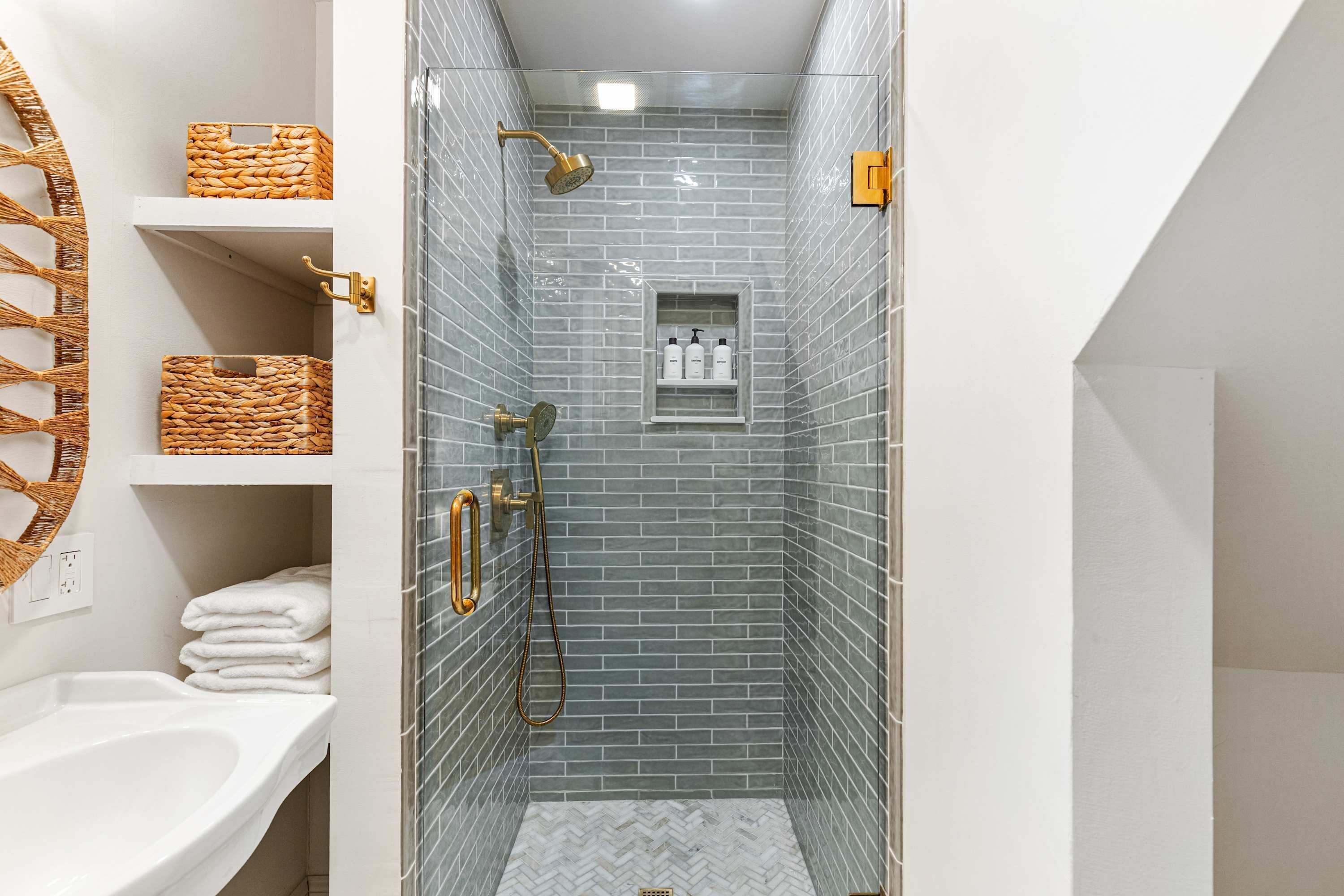 A narrow shower with a glass door, a white sink, and a towel rack with baskets.