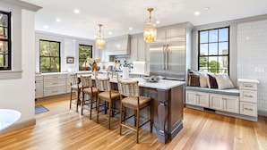 A modern kitchen with white cabinetry, a central island with bar stools, stainless steel appliances, and large windows. - The Royce on Pelham - Luxury Family Estate (Newport)