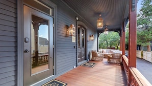 A covered porch with a wooden floor, a seating area with wicker furniture, and a door with a glass panel. - The Royce on Pelham - Luxury Family Estate (Newport)