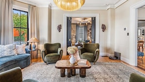 A living room with a sofa, two armchairs, a coffee table with a vase and book, a chandelier, and a view of the outside. - The Royce on Pelham - Luxury Family Estate (Newport)