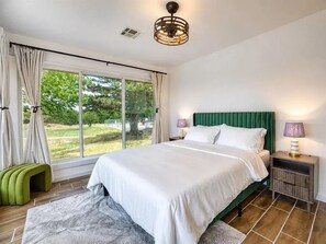 Makuuhuoneessa on suuri ikkuna, valkoisilla lakanoiden peitetty sänky, vihreä pääty ja yöpöytä, jossa on lamppu, sekä vihreä penkki.